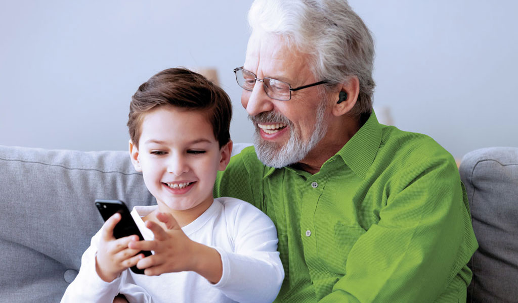 Opa sitzt mit Enkel auf dem Sofa und lässt sich das Handy zeigen. Imagebild für gutes hören.