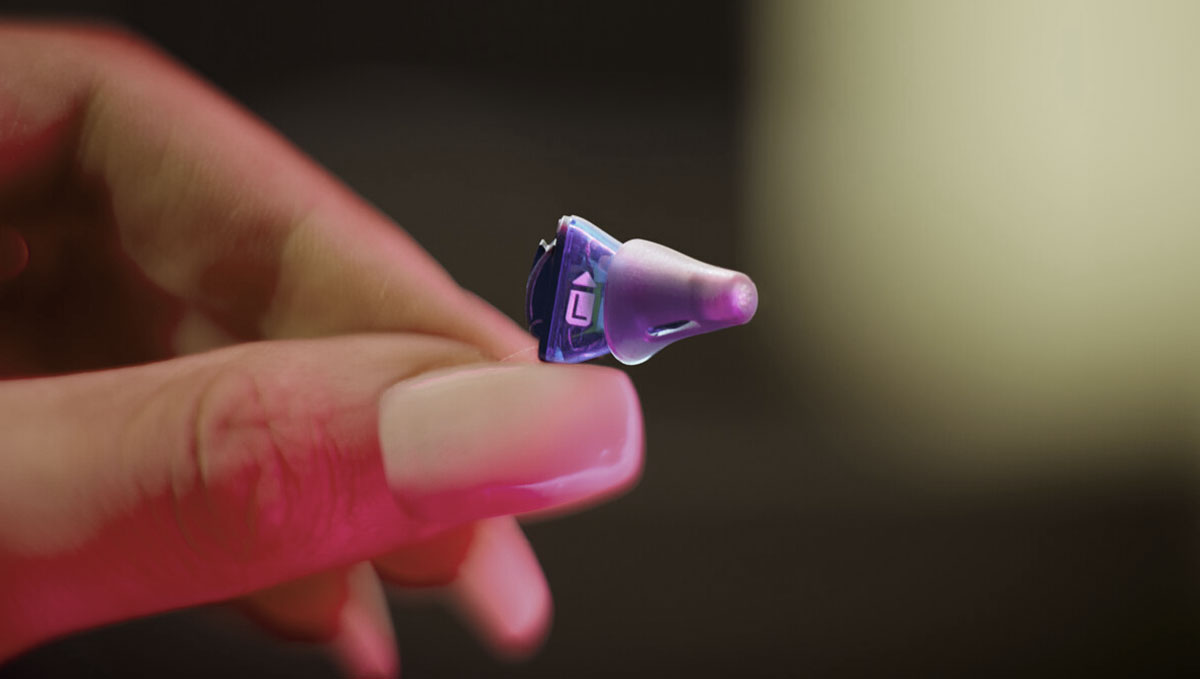 Detailansicht eines etwa 5 mal 10 mm kleines Hörgerät wird von zwei Fingern gehalten und präsentiert